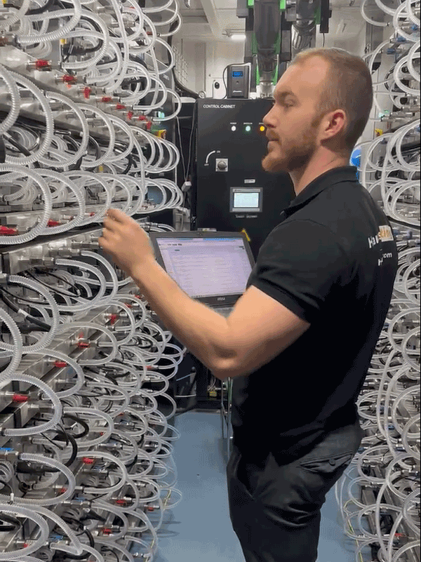 A side view of a professional wearing a Hamus Hosting shirt, plugging an internet cable into a miner inside a Bitmain HK3 hydro container, surrounded by high-performance mining equipment.