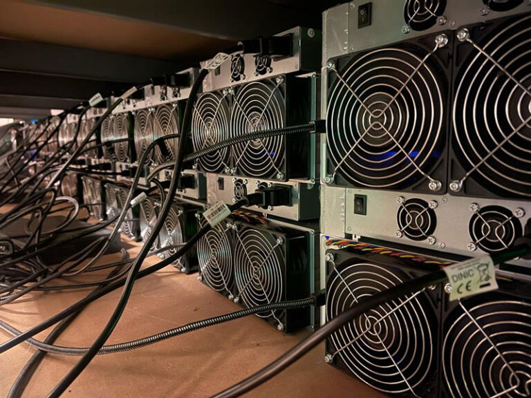 A row of ASIC miners stacked on shelves, connected with power cables, and actively cooling with large fans.